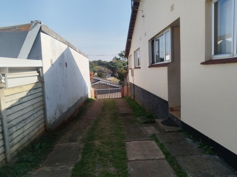 A side exterior view showing a paved driveway leading to a metal security gate, flanked by a residential building and a boundary wall.