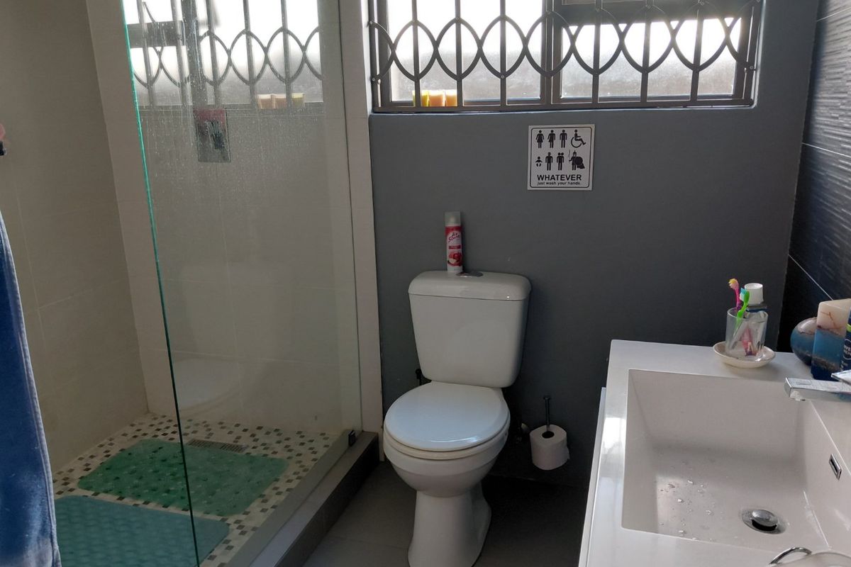 A functional bathroom featuring a glass-enclosed shower, a modern vanity basin, and a toilet, finished with tiled flooring and security bars on the window.
