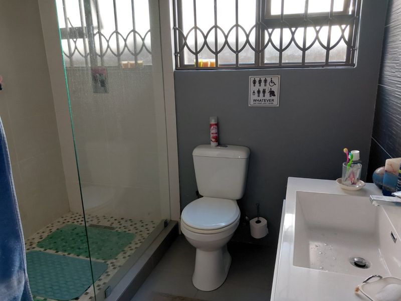 A functional bathroom featuring a glass-enclosed shower, a modern vanity basin, and a toilet, finished with tiled flooring and security bars on the window.