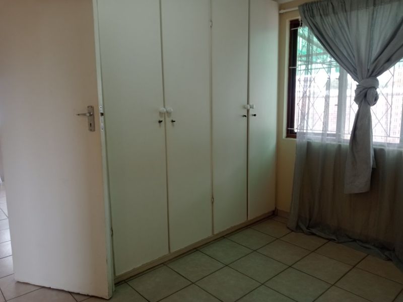 A bedroom featuring built-in cupboards, tiled flooring, and a window fitted with security bars.