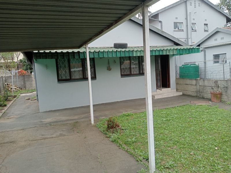 Rear exterior view of the apartment featuring a covered carport, concrete driveway, and a small private garden area with perimeter walling.