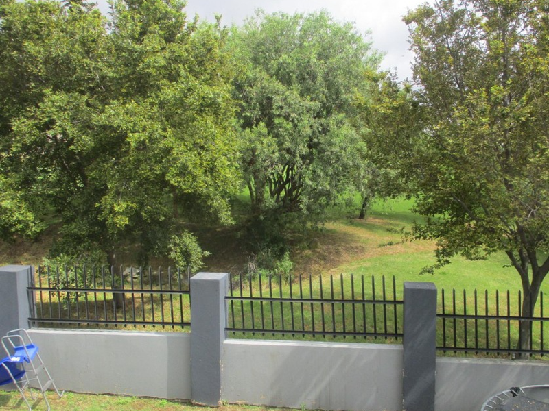 A view of the rear perimeter wall featuring concrete pillars and black metal security fencing, overlooking a lush green garden area with mature trees.