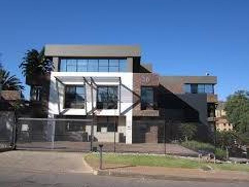 A modern commercial building facade featuring large glass windows, a paved driveway, and a secure metal gate entrance.