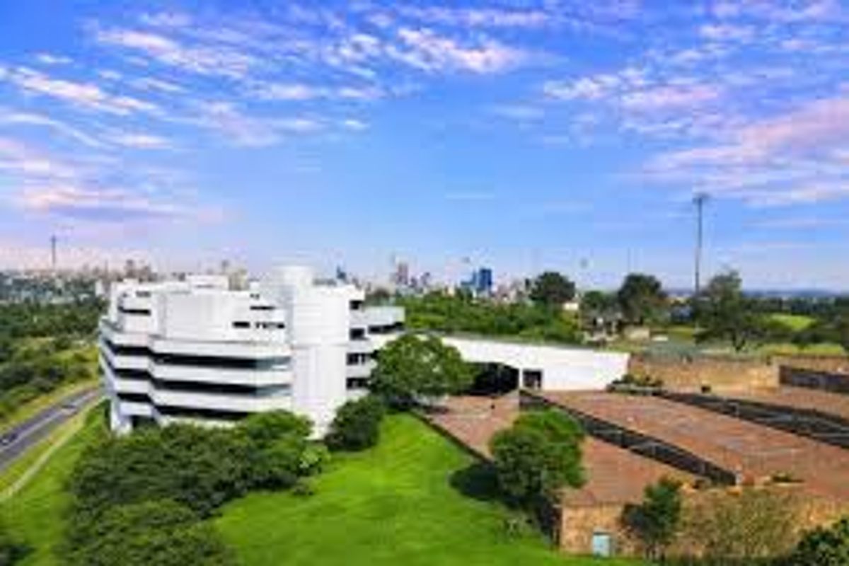Aerial view of a modern commercial office complex set within landscaped grounds, featuring paved parking areas and distant city skyline views.