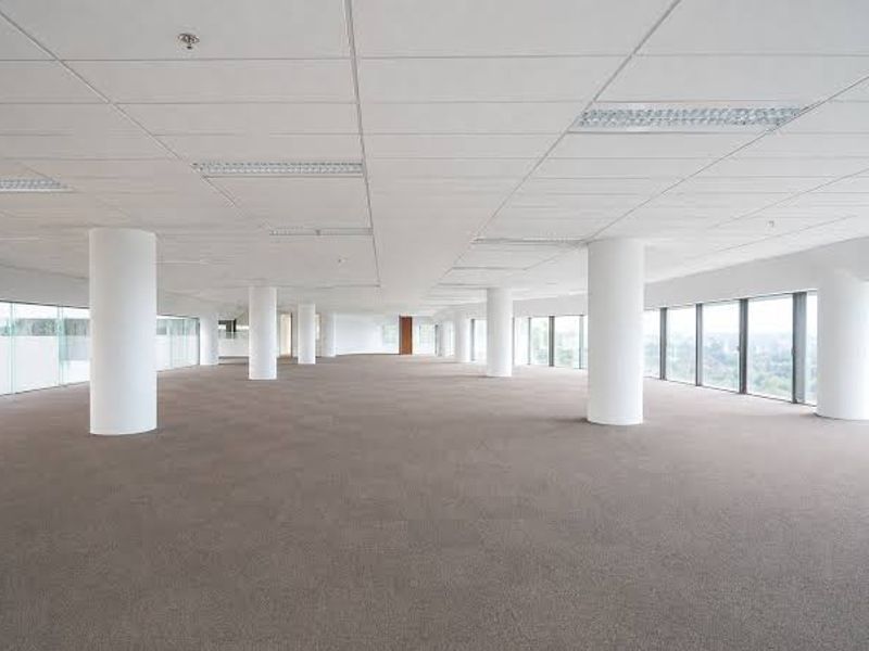 A spacious, open-plan commercial office area featuring large windows, structural columns, suspended ceilings, and neutral carpet tiles, offering a bright and versatile workspace.