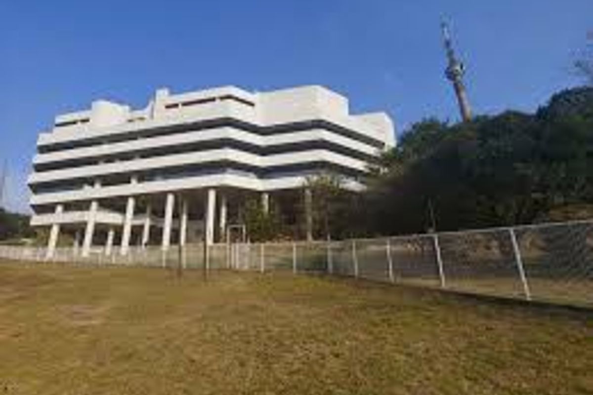 Exterior view of a multi-story commercial building featuring a white facade, concrete support pillars, and surrounding landscaped grounds with perimeter fencing.