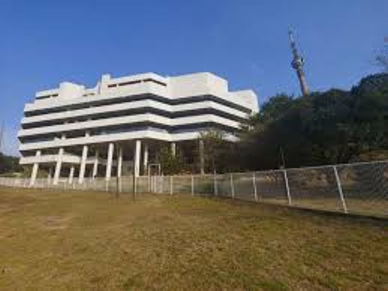 Exterior view of a multi-story commercial building featuring a white facade, concrete support pillars, and surrounding landscaped grounds with perimeter fencing.