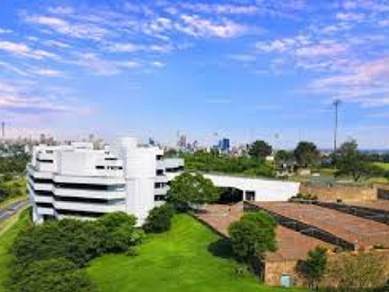 Aerial view of a modern commercial office complex set within landscaped grounds, featuring paved parking areas and distant city skyline views.