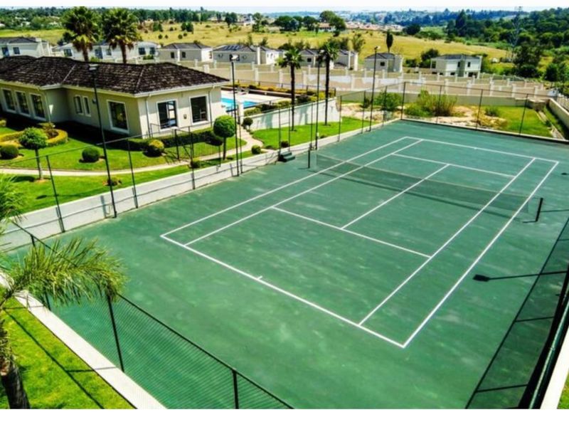 An elevated view of a well-maintained private tennis court with perimeter fencing and floodlights, situated within a residential estate.