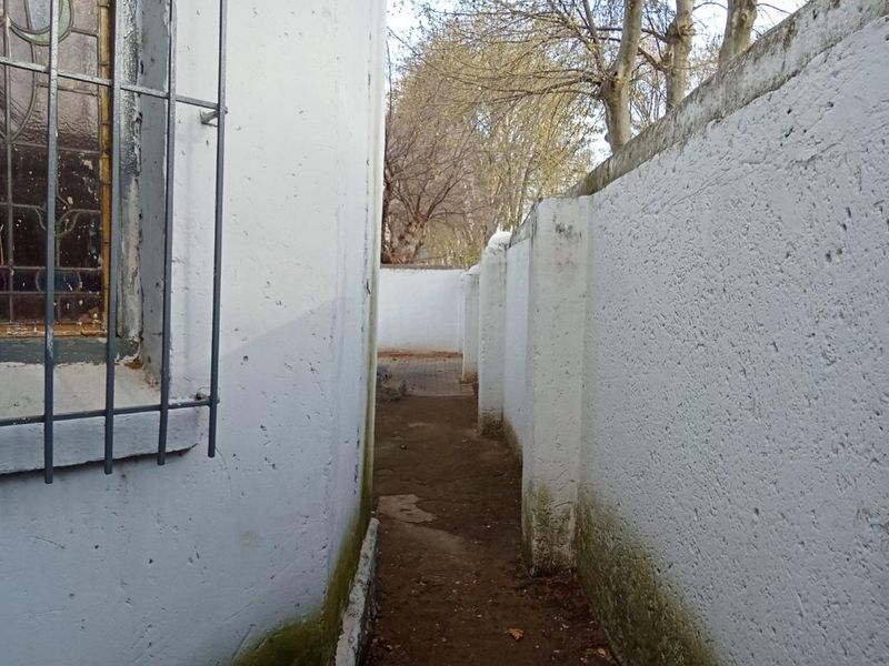 A narrow side passage between the house exterior and a high boundary wall, showing signs of weathering and damp at the base.
