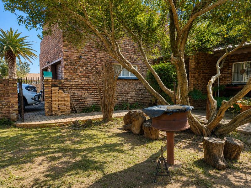 A rear garden area featuring a face brick facade, paved walkway, and a built-in braai area under mature trees.