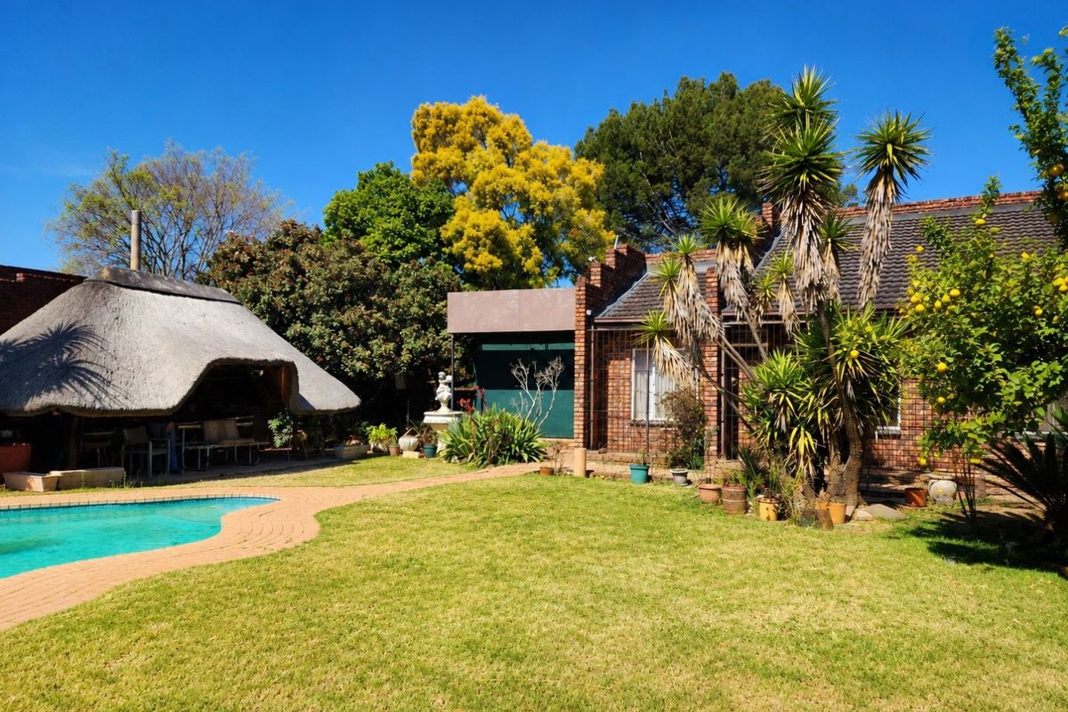 Rear garden view featuring a swimming pool, a traditional thatched lapa, and established landscaping with brick-built house exterior.