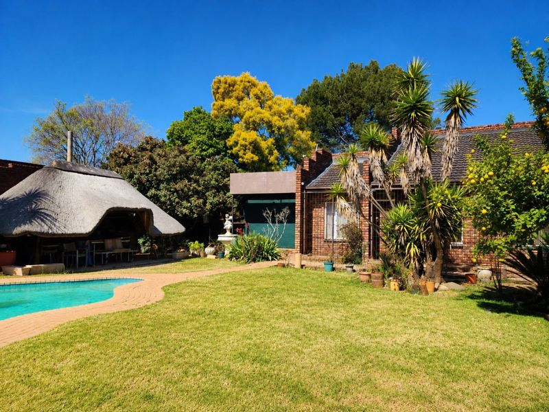 Rear garden view featuring a swimming pool, a traditional thatched lapa, and established landscaping with brick-built house exterior.