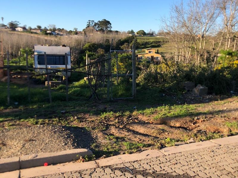 Street view of a vacant residential plot featuring a metal gate, wire fencing, and natural landscape with surrounding neighborhood views.