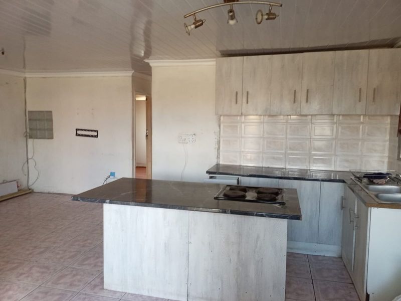 Open plan kitchen area featuring a central island with hob, built-in cabinetry, tiled backsplash, and tiled flooring in a neutral, dated aesthetic.