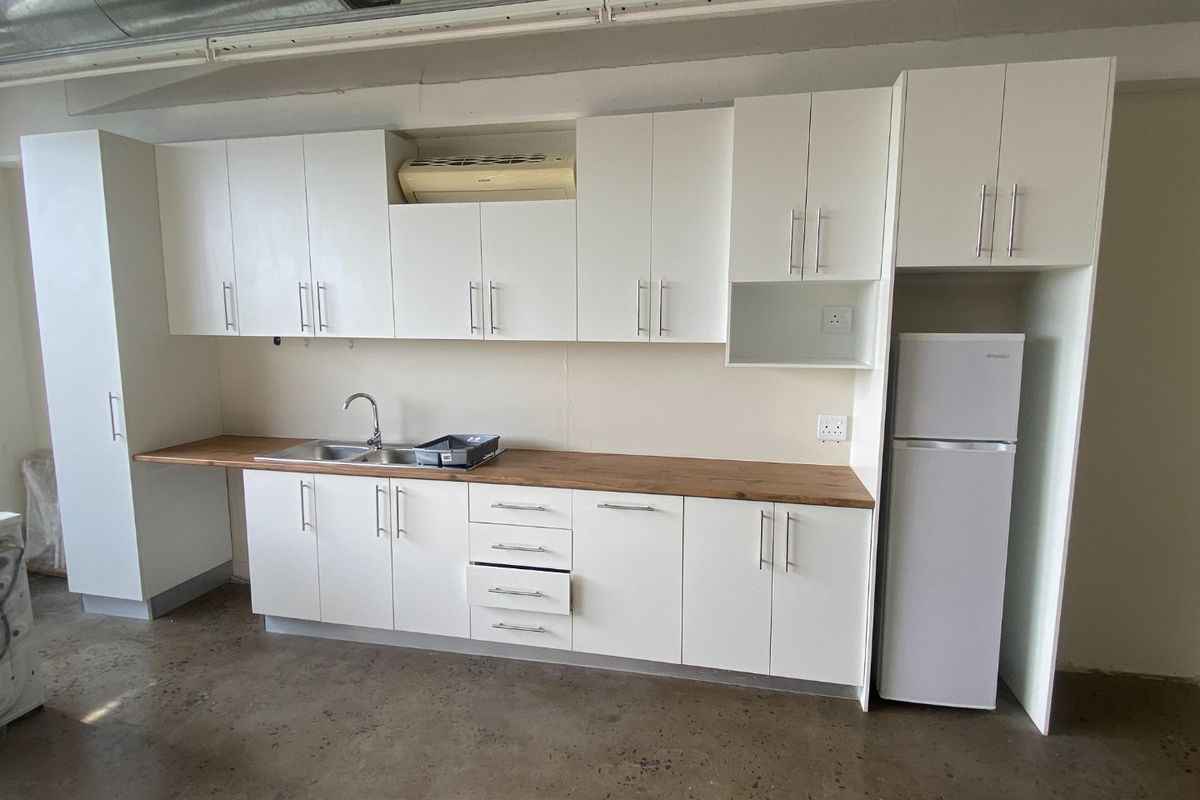 A functional kitchenette featuring white cabinetry, a wooden countertop, stainless steel sink, and exposed industrial-style ductwork in an open-plan commercial space.