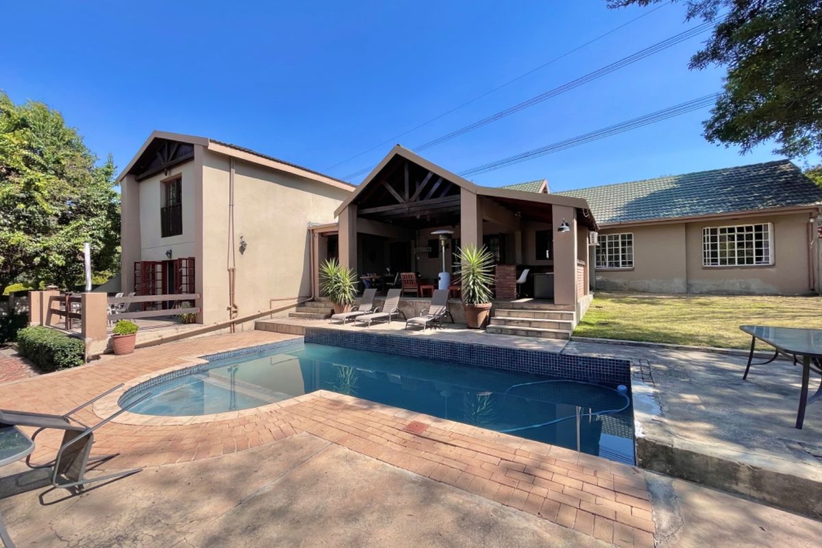 Rear exterior view featuring a swimming pool, paved surround, and a covered patio area with exposed timber trusses, set against a landscaped garden.