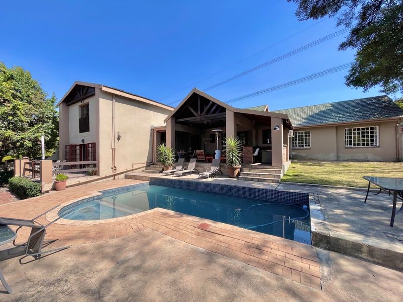 Rear exterior view featuring a swimming pool, paved surround, and a covered patio area with exposed timber trusses, set against a landscaped garden.