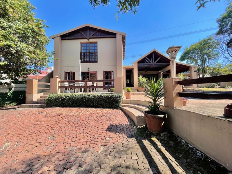 Rear view of the property featuring a paved patio area, timber balcony, and a cobblestone driveway leading to the main structure.