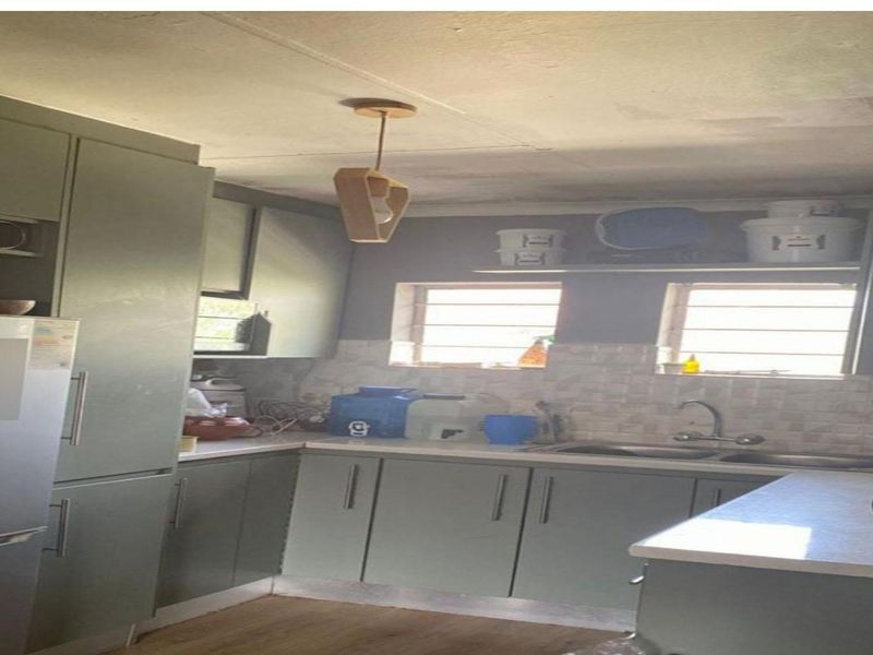 A compact kitchen featuring sage green cabinetry, laminate countertops, a double basin sink, and tiled backsplash, requiring some ceiling maintenance.