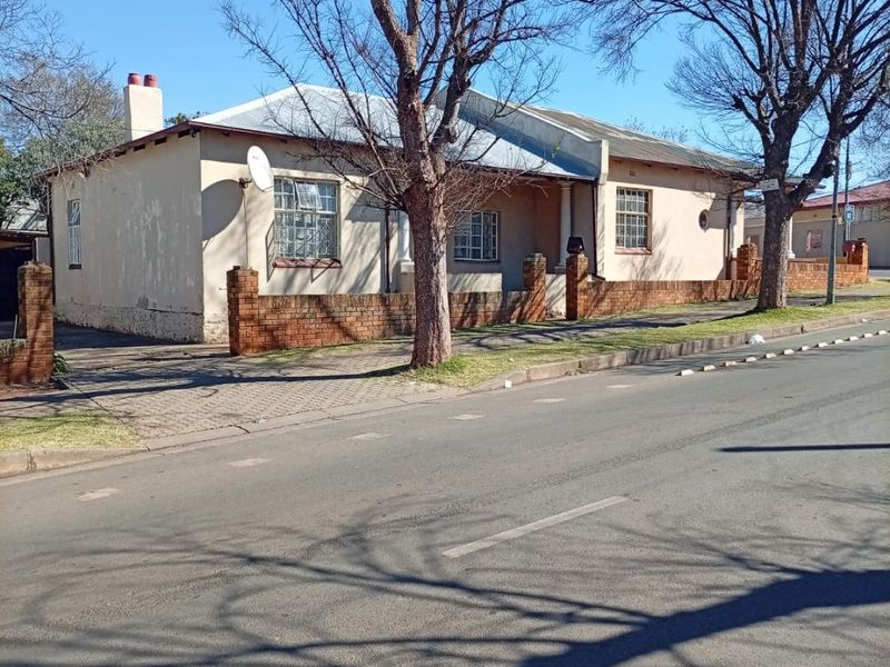 Front elevation of a single-story house featuring a brick boundary wall, paved driveway, and corrugated metal roof, showing signs of exterior wear.