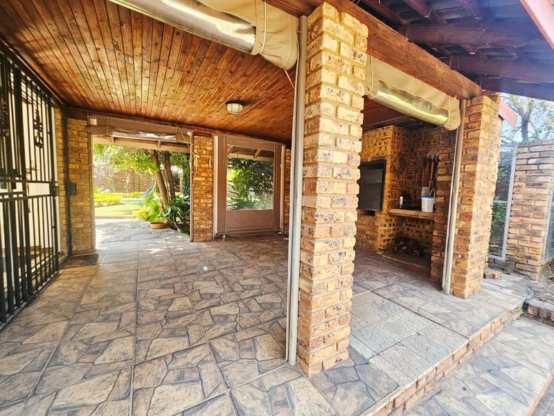 Covered outdoor patio featuring exposed brick pillars, a built-in braai, timber ceiling, and stone-look tiled flooring, perfect for alfresco entertaining.