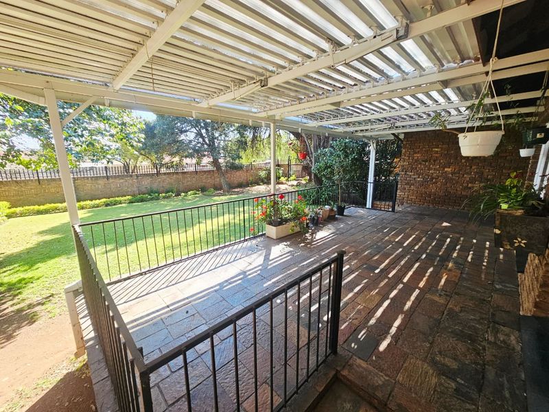 A covered outdoor patio featuring stone tile flooring, a metal pergola roof structure, and a metal railing overlooking a spacious garden.