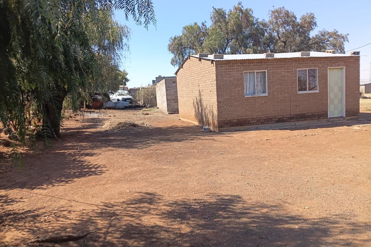 A view of the rear exterior of a simple brick house with a corrugated metal roof and a large, unpaved yard area.