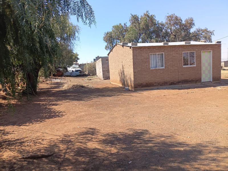 A view of the rear exterior of a simple brick house with a corrugated metal roof and a large, unpaved yard area.