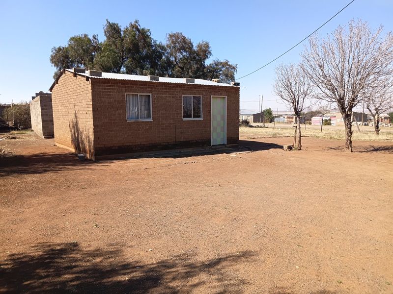 A detached single-story house featuring exposed brick walls and a corrugated metal roof, situated on a large, undeveloped dirt plot.