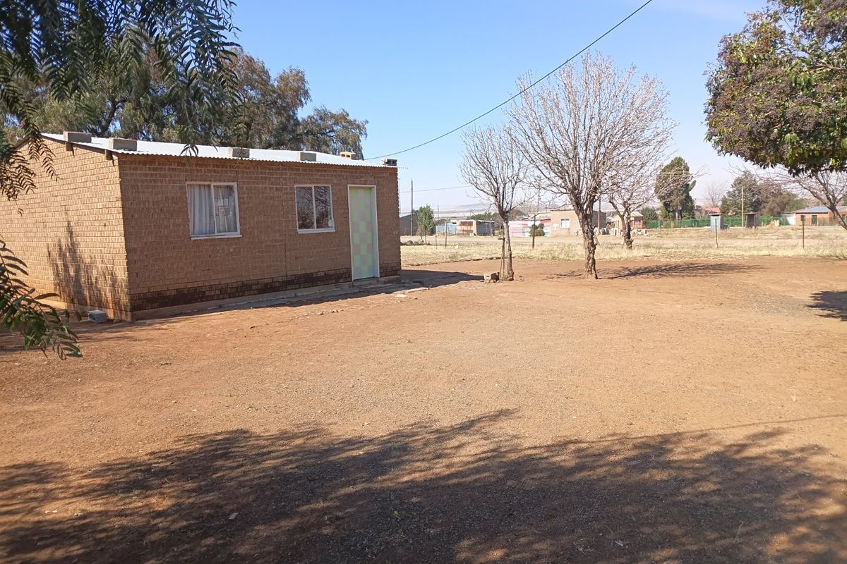 A view of the rear exterior of a single-story brick house with a corrugated metal roof and a large, unpaved yard area.