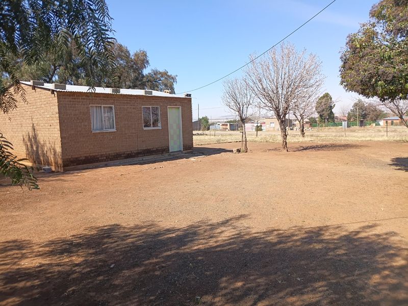 A view of the rear exterior of a single-story brick house with a corrugated metal roof and a large, unpaved yard area.