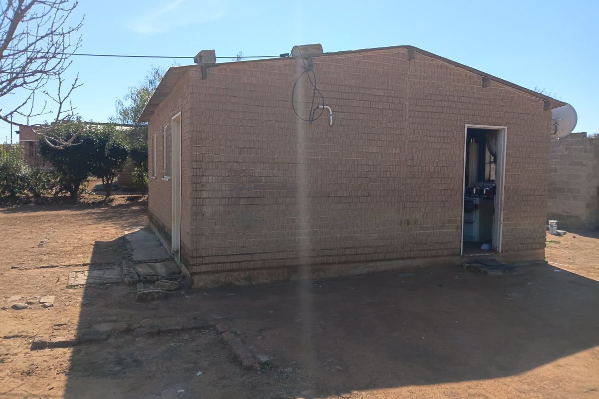 Rear view of a single-story brick house featuring exposed exterior wiring and a satellite dish, set on an unpaved plot.