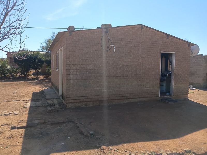 Rear view of a single-story brick house featuring exposed exterior wiring and a satellite dish, set on an unpaved plot.