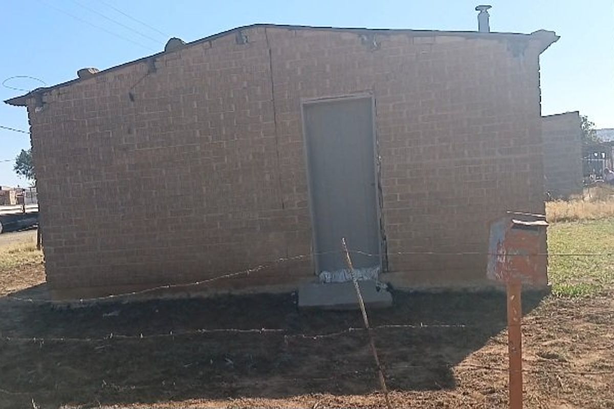 A single-story brick house exterior featuring a metal entrance door and a front yard enclosed by a barbed wire fence.