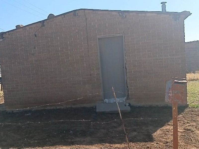 A single-story brick house exterior featuring a metal entrance door and a front yard enclosed by a barbed wire fence.
