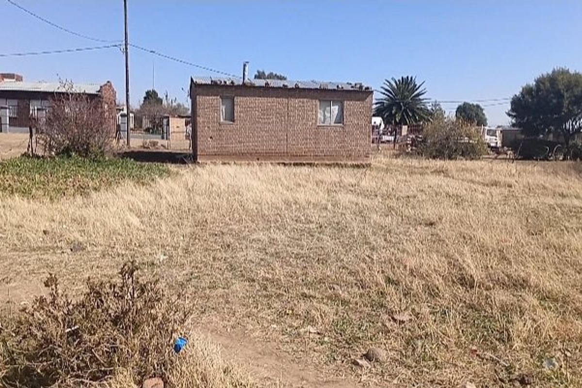 A view of a detached brick structure with a corrugated metal roof situated on a large, undeveloped plot of land.