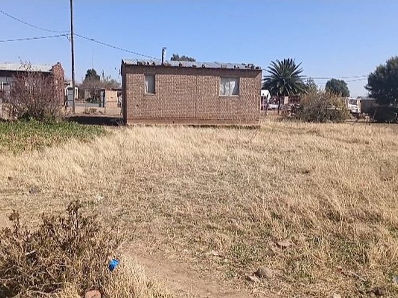A view of a detached brick structure with a corrugated metal roof situated on a large, undeveloped plot of land.