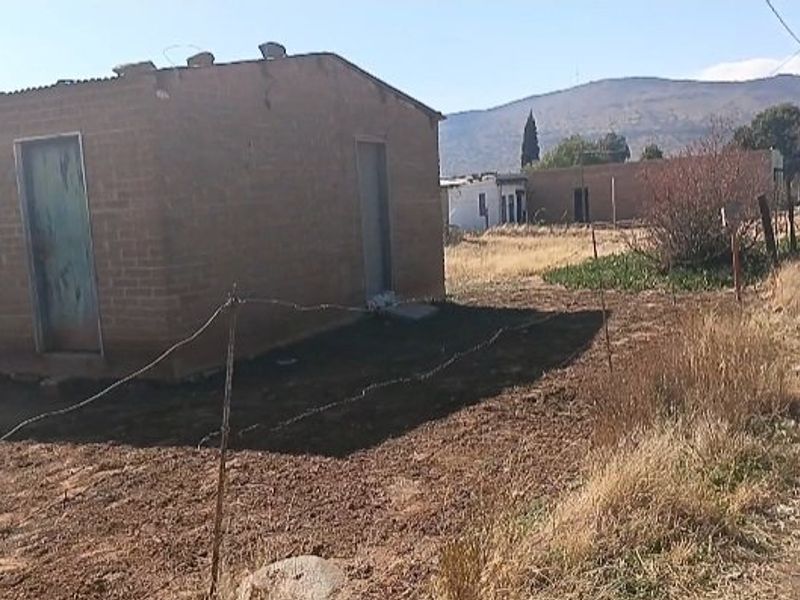 A basic detached brick structure with a corrugated metal roof, situated on a rural plot with wire fencing and mountain views.
