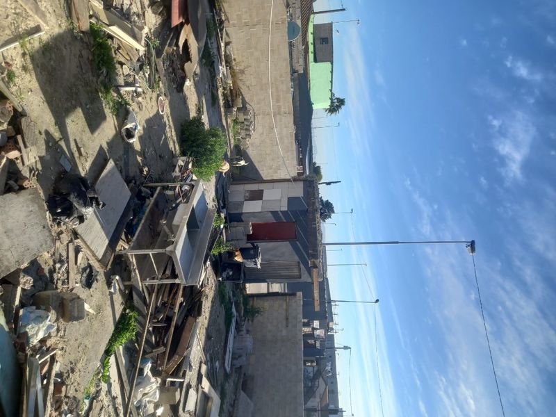 A vacant residential plot featuring a concrete block boundary wall, a small shed structure, and a metal utility sink amidst significant site debris.