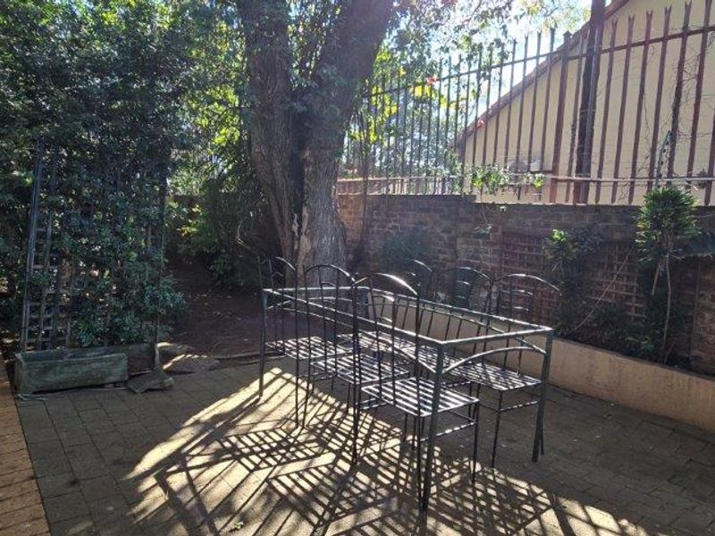 A paved outdoor patio area featuring a retaining wall and perimeter fencing, surrounded by mature trees and garden greenery.