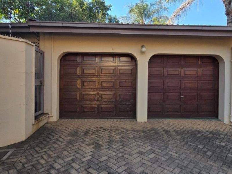 A double garage with wooden panel doors and paved driveway access, featuring exterior security fencing and wall-mounted lighting.