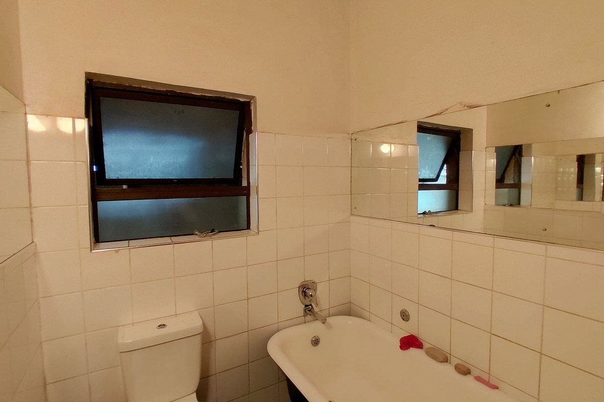 A dated bathroom featuring white wall tiles, a standard bathtub, a toilet, and a wall-mounted mirror, requiring cosmetic updates to modernize the space.