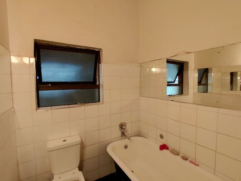 A dated bathroom featuring white wall tiles, a standard bathtub, a toilet, and a wall-mounted mirror, requiring cosmetic updates to modernize the space.