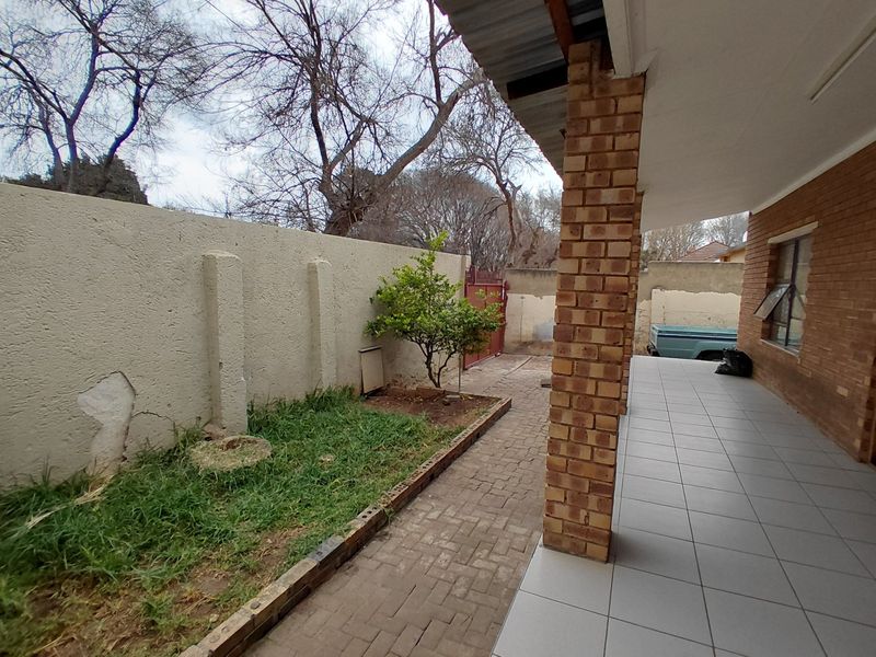 A covered patio area featuring tiled flooring and a brick pillar, overlooking a small garden space with a paved walkway and boundary wall.