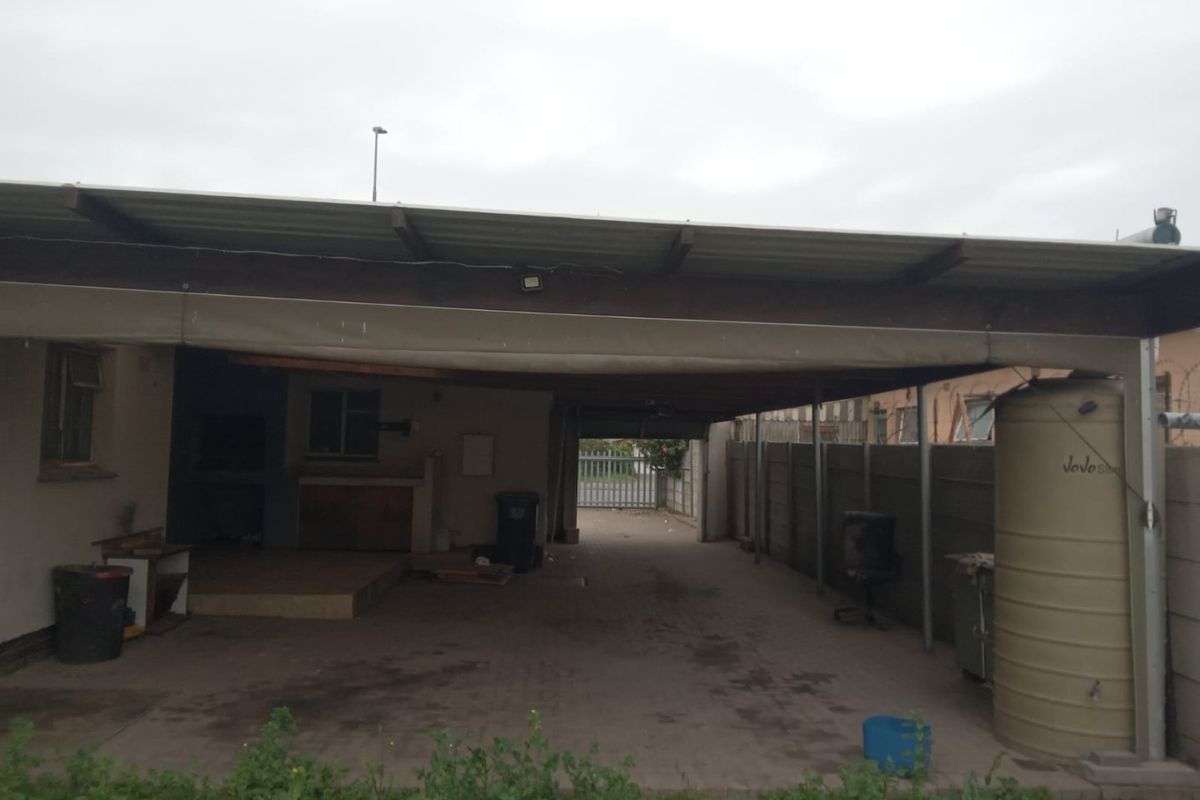 Rear exterior area featuring a large covered patio, paved driveway, built-in braai, and a water storage tank enclosed by precast concrete walling.