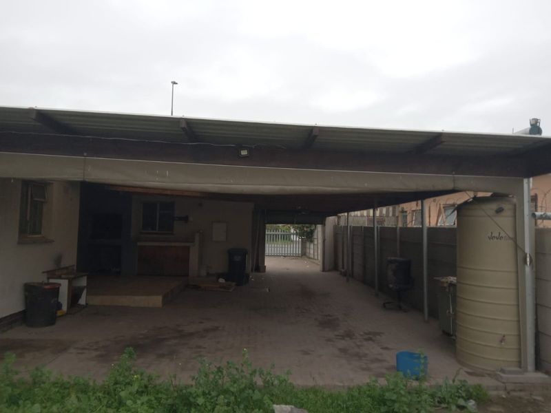 Rear exterior area featuring a large covered patio, paved driveway, built-in braai, and a water storage tank enclosed by precast concrete walling.