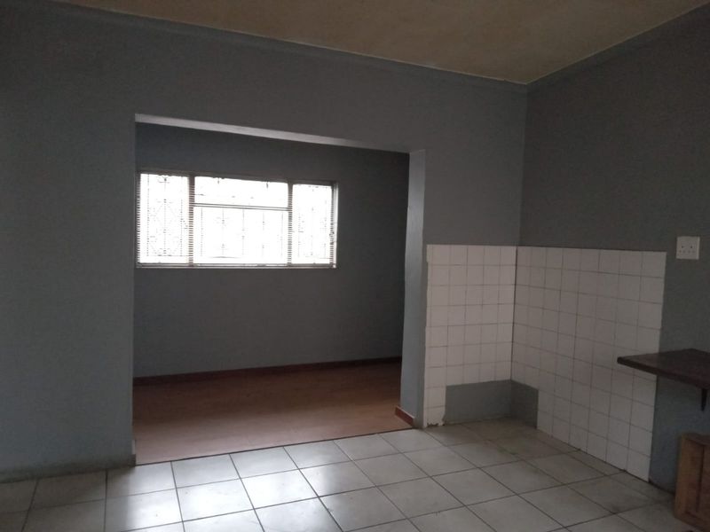 An empty open-plan area featuring a combination of tiled and laminate flooring with a tiled kitchen splashback and security bars on the window.