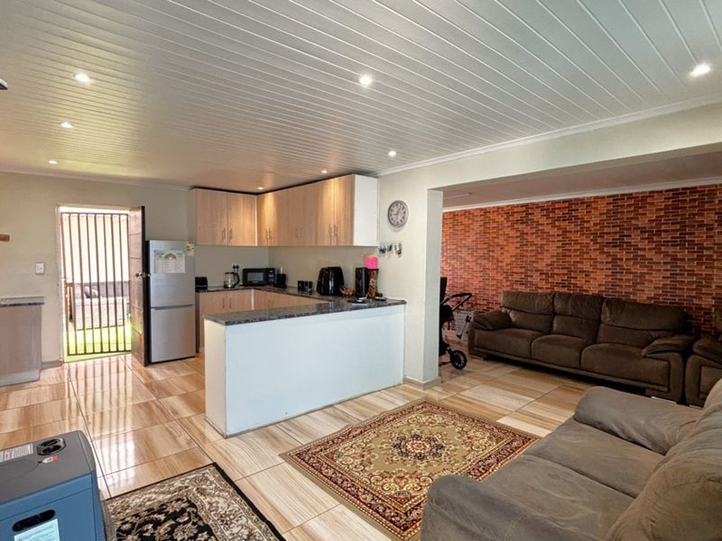 Open plan living area featuring tiled floors, a tongue and groove ceiling, and a kitchen with granite countertops and a brick feature wall.