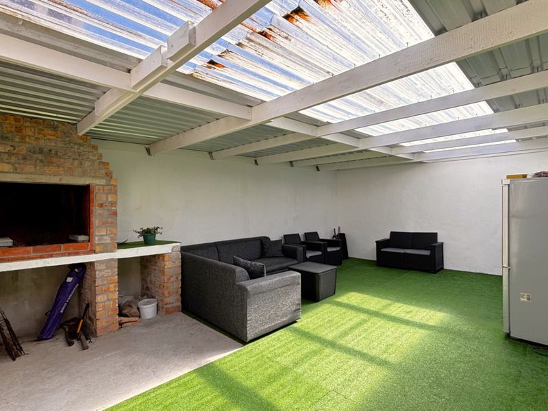 Covered outdoor patio area featuring a built-in brick braai, timber beam structure with translucent roofing, and artificial grass flooring.